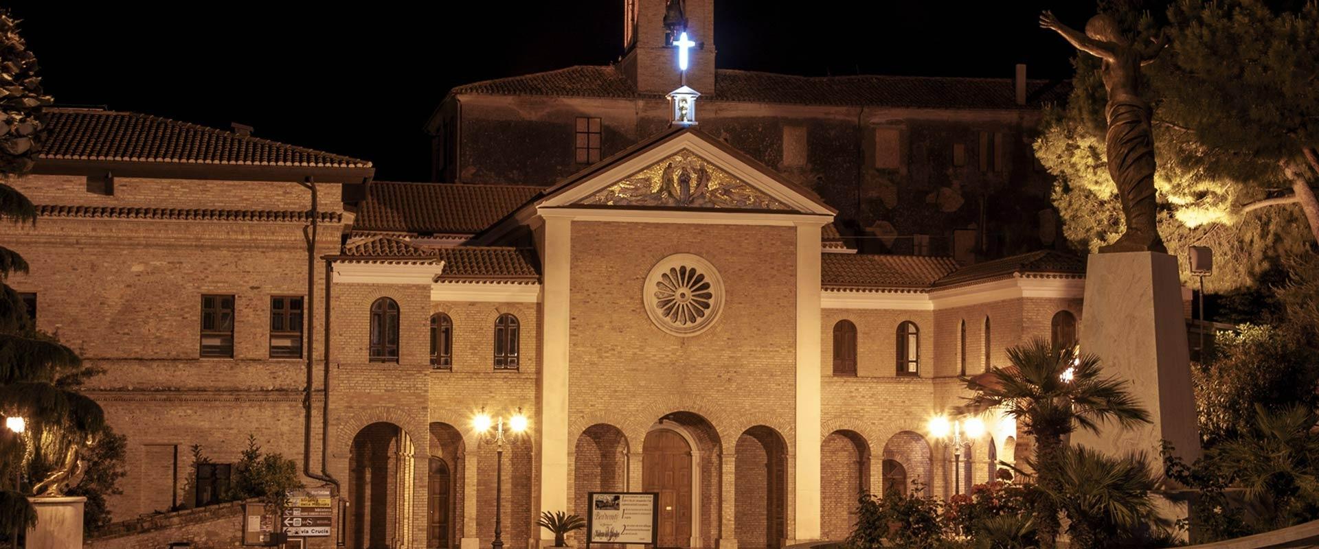 Chiesa illuminata di notte, atmosfera serena con statua in primo piano e croce luminosa.
