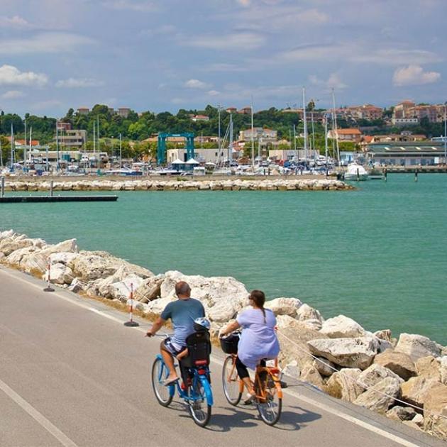 Due ciclisti pedalano lungo la costa, mare turchese e città in lontananza.