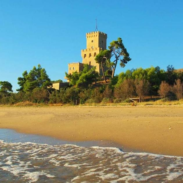 Torre medievale su collina verde, spiaggia dorata e mare calmo sotto cielo azzurro.