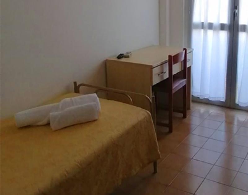 Stanza semplice e luminosa, letto singolo, scrivania, sedia e balcone con tende chiare.